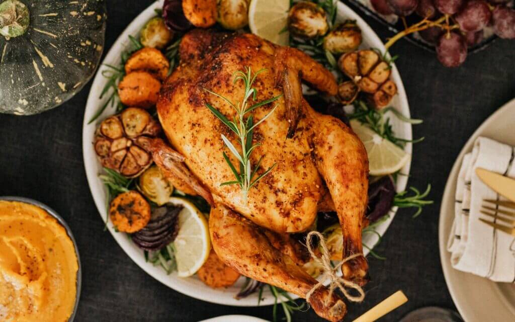 A roasted whole chicken garnished with fresh rosemary, sliced lemons, and surrounded by roasted vegetables such as Brussels sprouts, sweet potatoes, and garlic cloves, is displayed on a platter. Nearby, a pumpkin and a bunch of grapes are visible.