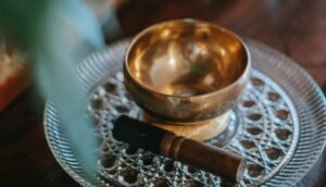 A brass singing bowl is placed on an ornate glass dish with an intricate pattern. Beside the bowl, there is a wooden mallet resting on the dish. The background is blurred with a hint of greenery visible on the left side.