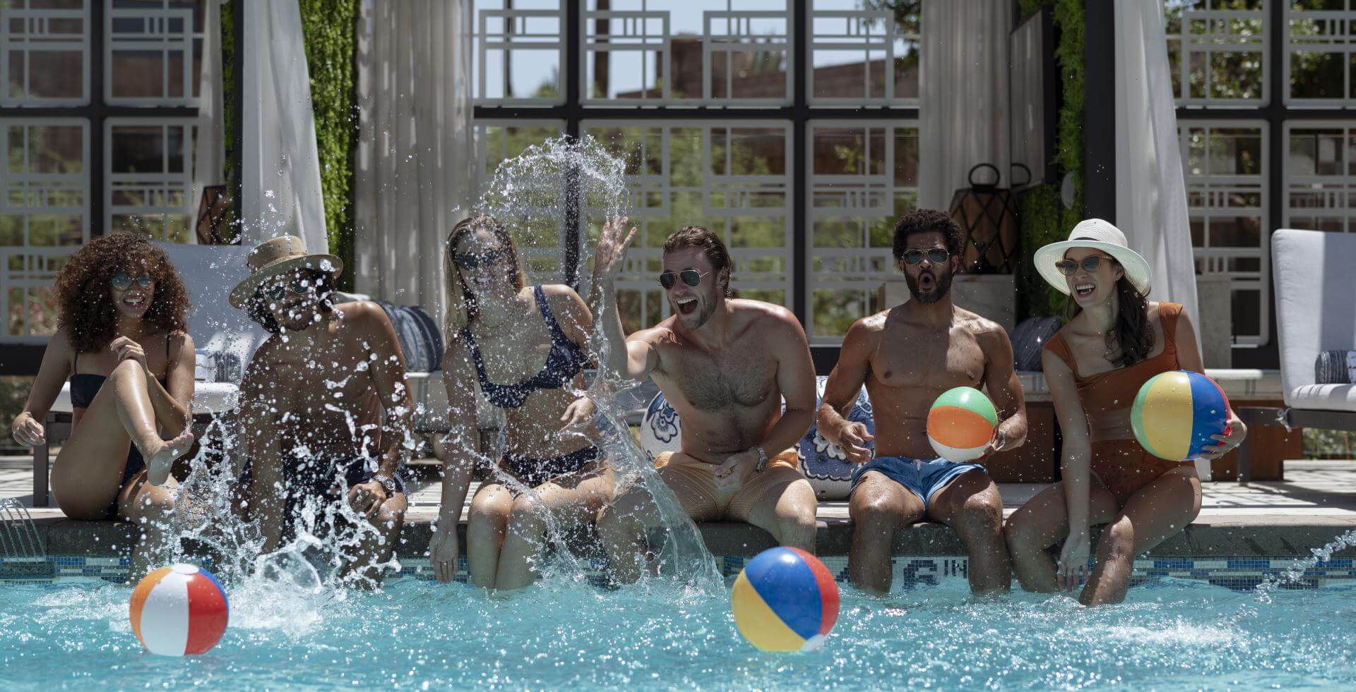 A group of six people, sitting by a pool, splash water and play with colorful beach balls. They are enjoying a sunny day, wearing swimsuits, and showing expressions of joy and excitement. The background features a stylish structure with modern design elements.