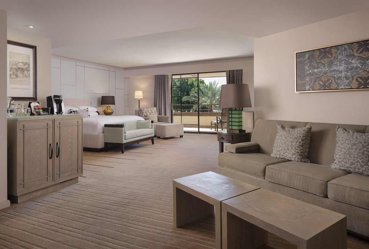 Spacious junior suite with a king-sized bed, a sofa set, a coffee station atop a cabinet, and a small wooden table. The room features a flat-screen TV, artwork on the walls, and a large sliding glass door leading to a balcony with views of greenery.
