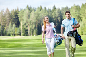A man and a woman walk on a golf course, smiling and carrying golf bags. They are dressed in casual golf attire, with the woman in a pink polo and white pants, and the man in a light blue polo and beige pants. Trees and green grass of the pristine golf course are visible in the background.