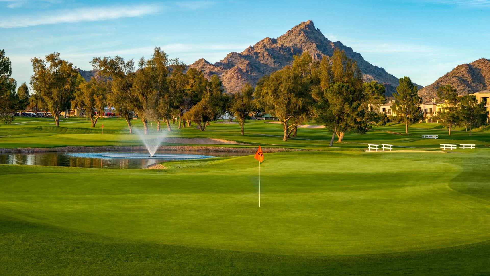 A picturesque golf course with a green field, a red flag marking a hole, and a pond with a water fountain. Tall trees surround the course, and a mountain range rises in the background under a clear blue sky.
