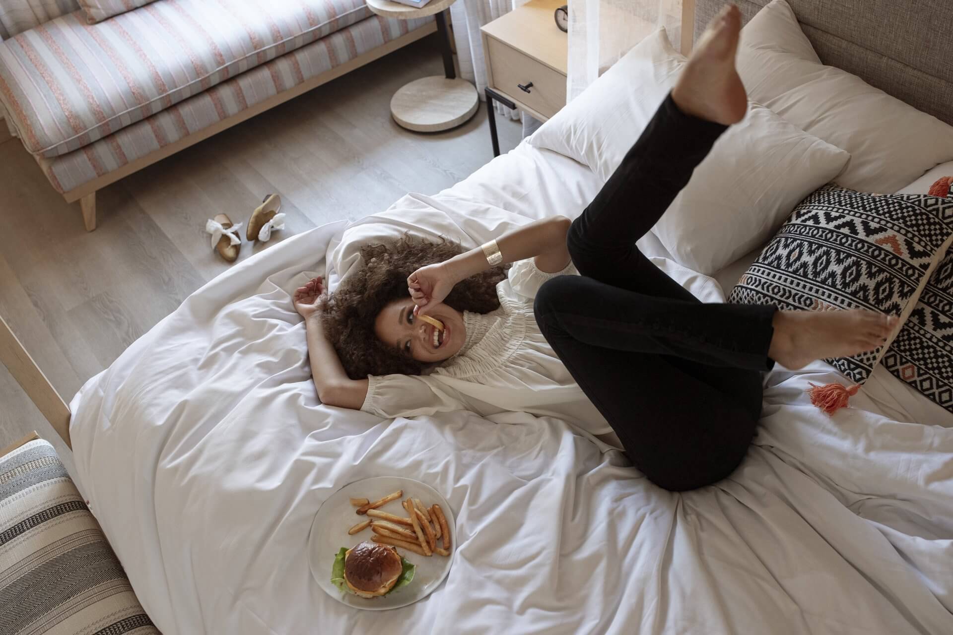 A person with curly hair playfully lies on a bed at the Arizona Biltmore, lifting one leg in the air while holding a french fry in their mouth. Next to them is a plate with a burger and fries. The room has a relaxed, cozy vibe with a couch, pillows, and shoes in the background.