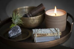 A tray holds a brass bowl with a wooden mallet, a lit candle in a ceramic holder, a green air plant on a black rock, and a bound bundle of sage labeled "TIERRA LUNA." The scene exudes a calming and zen-like atmosphere, perfect for any spa & wellness retreat.