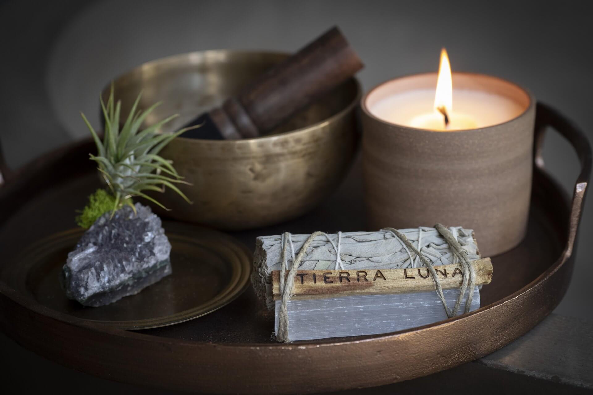 A tray holds a brass bowl with a wooden mallet, a lit candle in a ceramic holder, a green air plant on a black rock, and a bound bundle of sage labeled "TIERRA LUNA." The scene exudes a calming and zen-like atmosphere, perfect for any spa & wellness retreat.