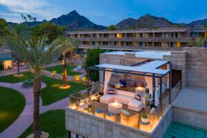 A rooftop lounge area with modern seating, fire pits, and light features atop a luxury building at the Arizona Biltmore, set against a backdrop of scenic mountains and lush greenery. Below, a courtyard with well-manicured lawns, palm trees, and additional outdoor seating is visible.