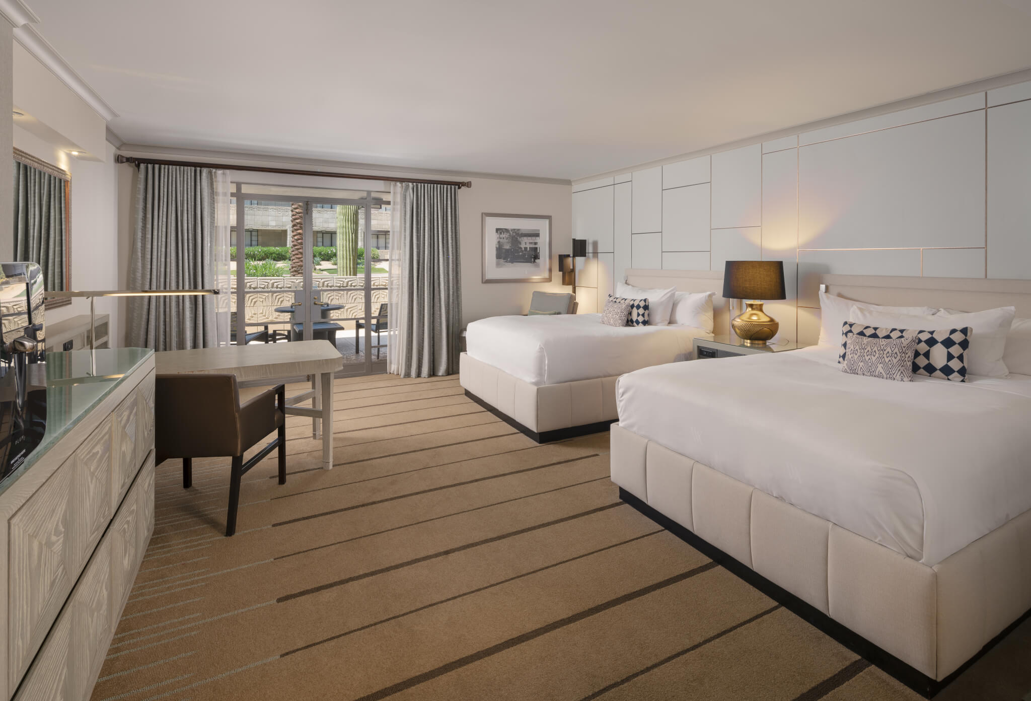 A spacious hotel room with two large beds, a desk and chair, a lamp, and neutral-colored decor. A glass door opens to a balcony with outdoor seating. A flat-screen TV is mounted on a light-colored cabinet along one wall. Walls are decorated with framed art.