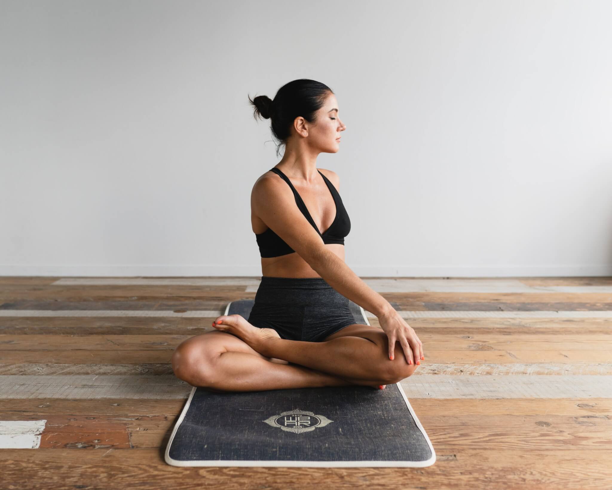 A woman with dark hair in a bun is sitting cross-legged on a yoga mat, performing a seated twist stretch. She is wearing a black sports bra and dark gray shorts. The setting appears to be a minimalist room with wooden flooring and a light-colored wall.