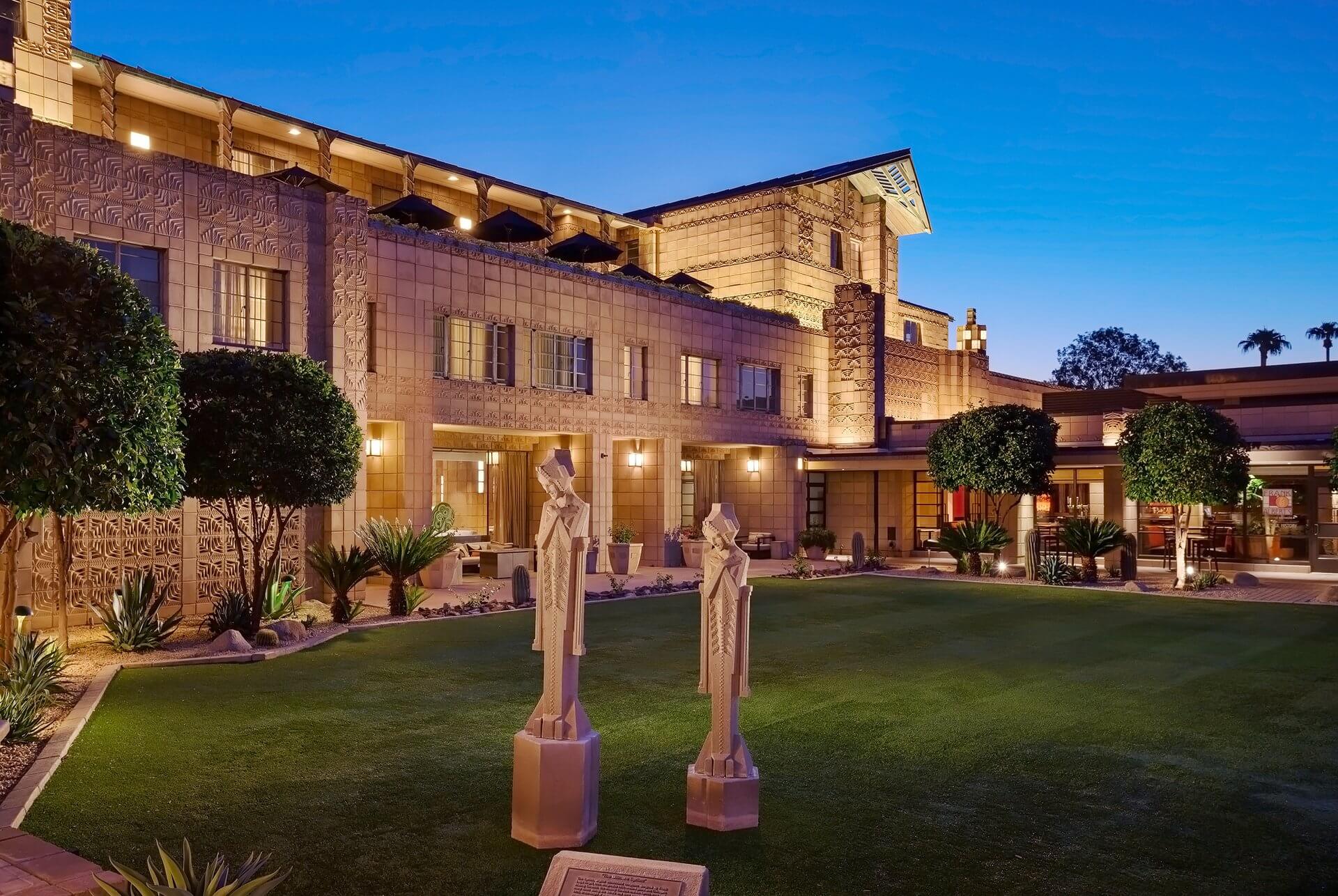 A large building with intricate patterns is lit up in the evening, embodying a reinvention of classic architecture. The foreground features a well-manicured lawn with two sculptures of human figures. Trimmed trees and shrubs line the sides, enhancing the tidy appearance of the landscape.