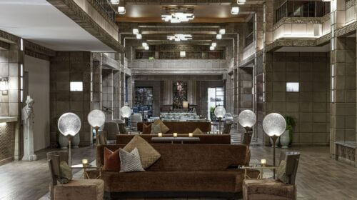 A spacious, elegant hotel lobby with modern decor. It features a brown couch with several cushions, wooden armchairs, and globe lamps on side tables. The room has high ceilings, intricate lighting fixtures, tiled walls, and a statue on the left side.