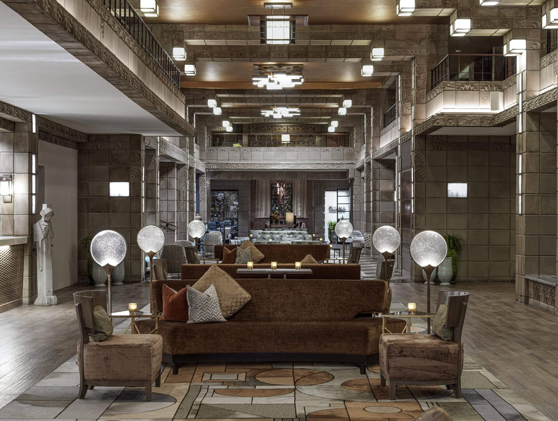 A spacious, elegant hotel lobby with modern decor. It features a brown couch with several cushions, wooden armchairs, and globe lamps on side tables. The room has high ceilings, intricate lighting fixtures, tiled walls, and a statue on the left side.