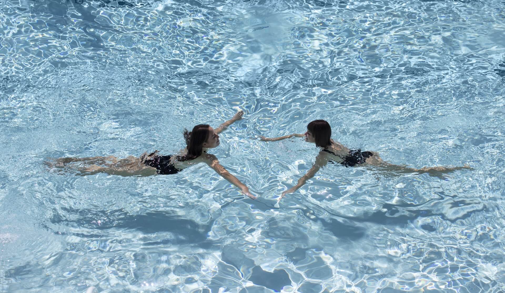 Two individuals are swimming towards each other in a clear blue resort pool. They are both wearing dark swimsuits and are reaching out their hands as they swim. The scene captures the reflections and movements of the water around them, creating a mesmerizing display in this idyllic setting.
