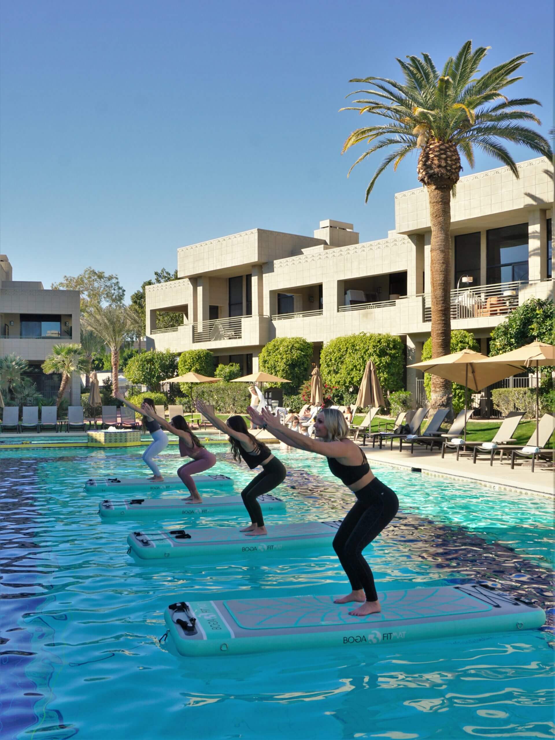 Four individuals are balancing on separate paddleboards in a pool, performing yoga poses with their arms extended forward and knees slightly bent. The serene spa & wellness setting is complete with lounge chairs, sun umbrellas, palm trees, and modern buildings in the background.