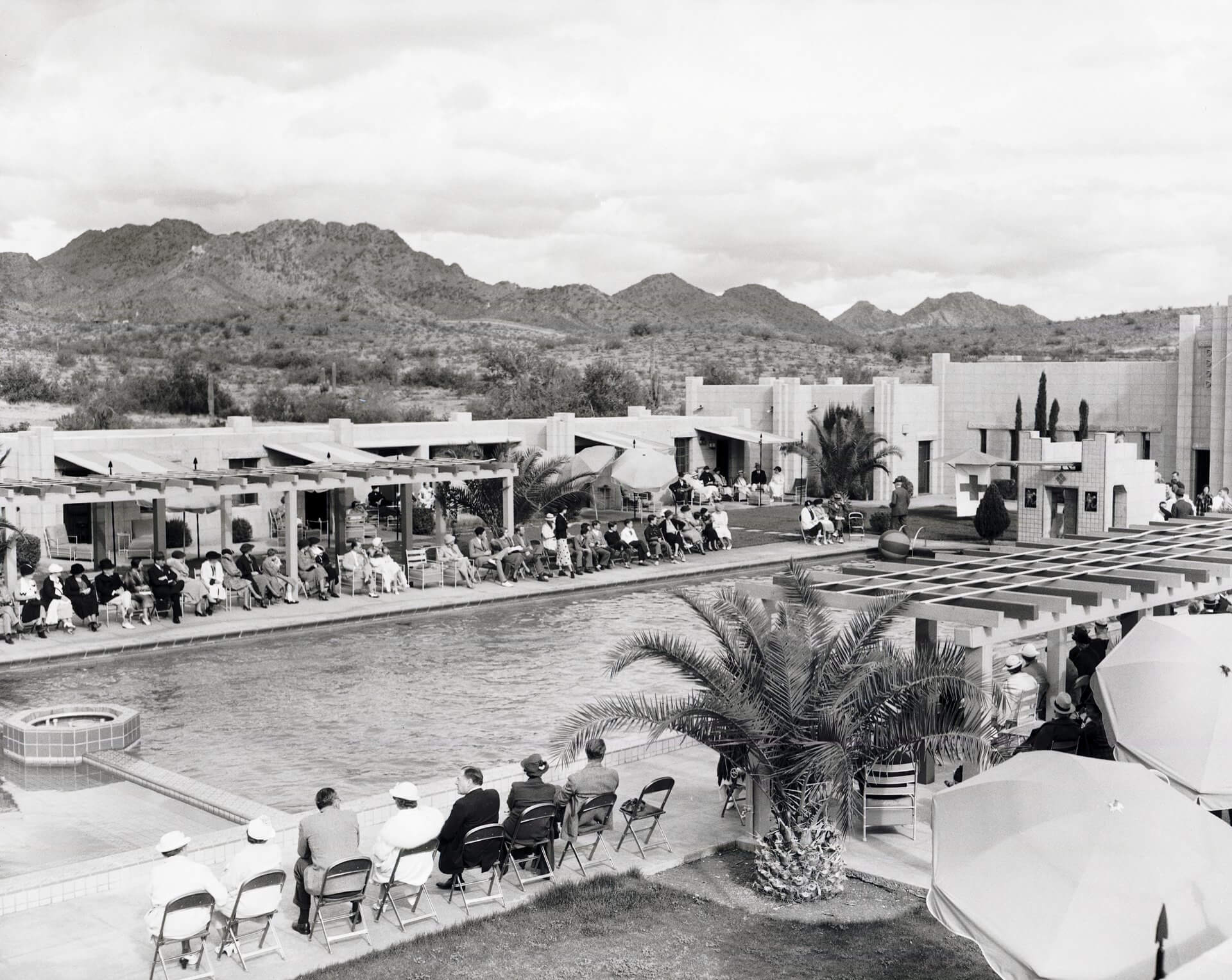 A mid-century outdoor scene featuring people seated around a large swimming pool in front of modern desert architecture, with mountains and an expansive sky in the background. Large umbrellas and poolside chairs are scattered about the foreground.