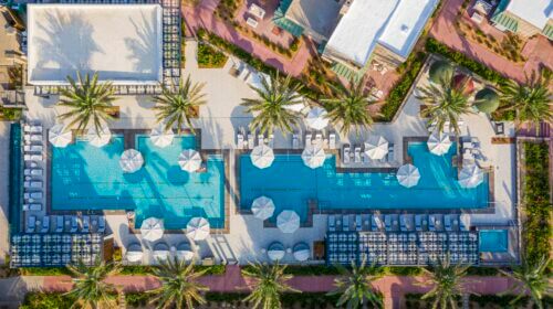 Aerial view of the luxurious Arizona Biltmore Pools at a resort. The scene features two large rectangular pools surrounded by palm trees, lounge chairs, and umbrellas. Adjacent buildings with white roofs and landscaped walkways are visible in the background.