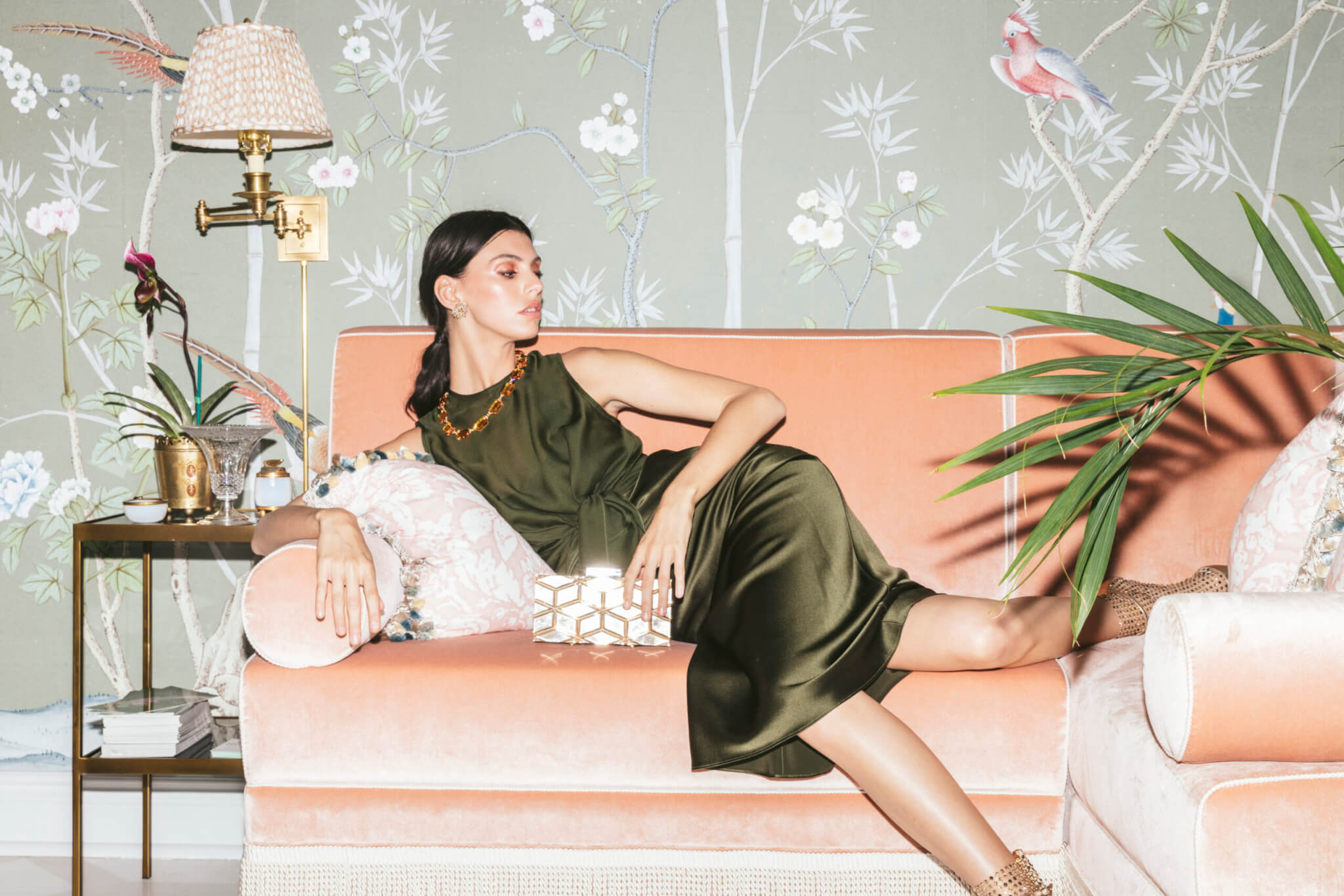 A woman in a green dress lounges on a peach velvet sofa next to a side table with a decorative lamp and a parrot statuette. The background, inspired by retail design trends, features floral and bird wallpaper. She holds a geometric clutch with gold accents, her gaze directed away.