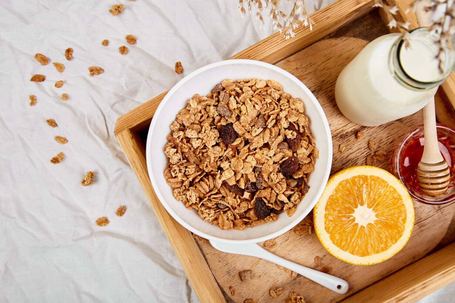 A bowl of granola with nuts and dried fruits sits on a wooden tray next to a bottle of milk, an orange half, and a small jar of honey with a dipper. Scattered granola pieces and a white cloth provide a cozy bed & breakfast ambiance.