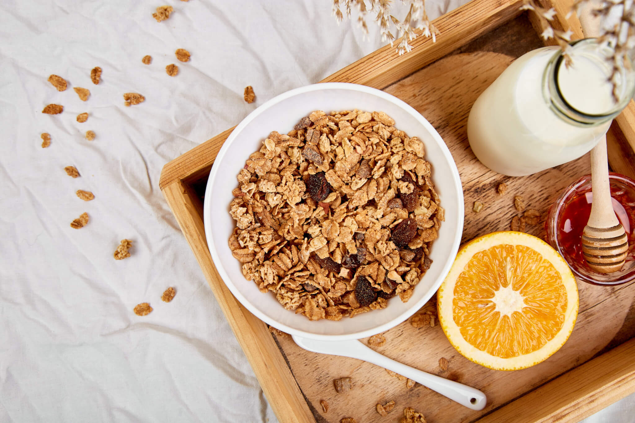A bowl of granola with nuts and dried fruits sits on a wooden tray next to a bottle of milk, an orange half, and a small jar of honey with a dipper. Scattered granola pieces and a white cloth provide a cozy bed & breakfast ambiance.