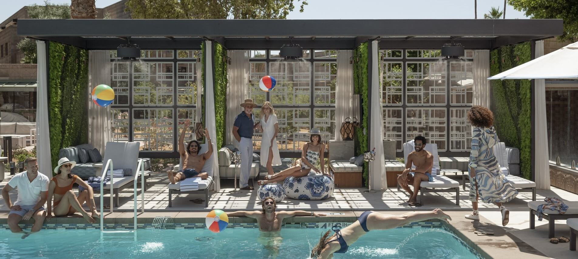 A group of people relax by a luxury pool surrounded by greenery and stylish decor. Some lounge on sunbeds while others stand, talking and playing with beach balls. One person dives into the pool, and the scene is framed by a modern pergola structure.