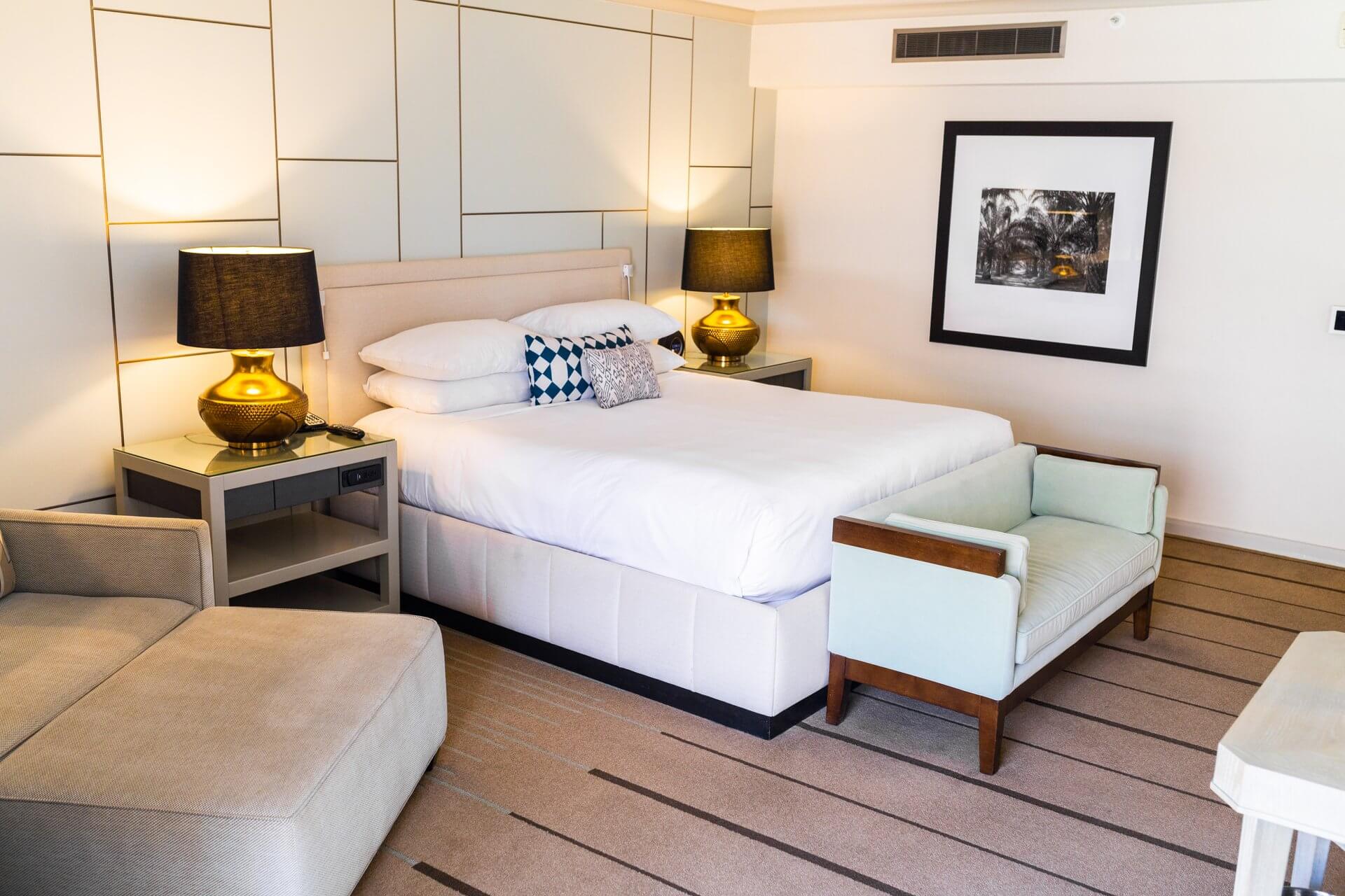 A modern Citrus Club balcony room features a large, neatly made bed with white linens, flanked by two nightstands with matching lamps. A small sofa bench sits at the foot of the bed. A beige armchair is positioned in the corner, and a framed artwork hangs above the bed.