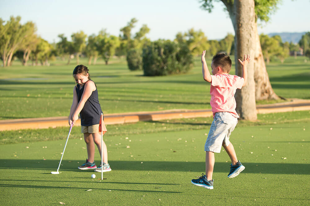 Two children are shown on a golf course. A young girl is about to putt a golf ball with a putter, wearing a dark sleeveless top and beige shorts. A young boy in a pink shirt and khaki shorts is joyfully celebrating with arms raised beside her, capturing the spirit of golf fun.