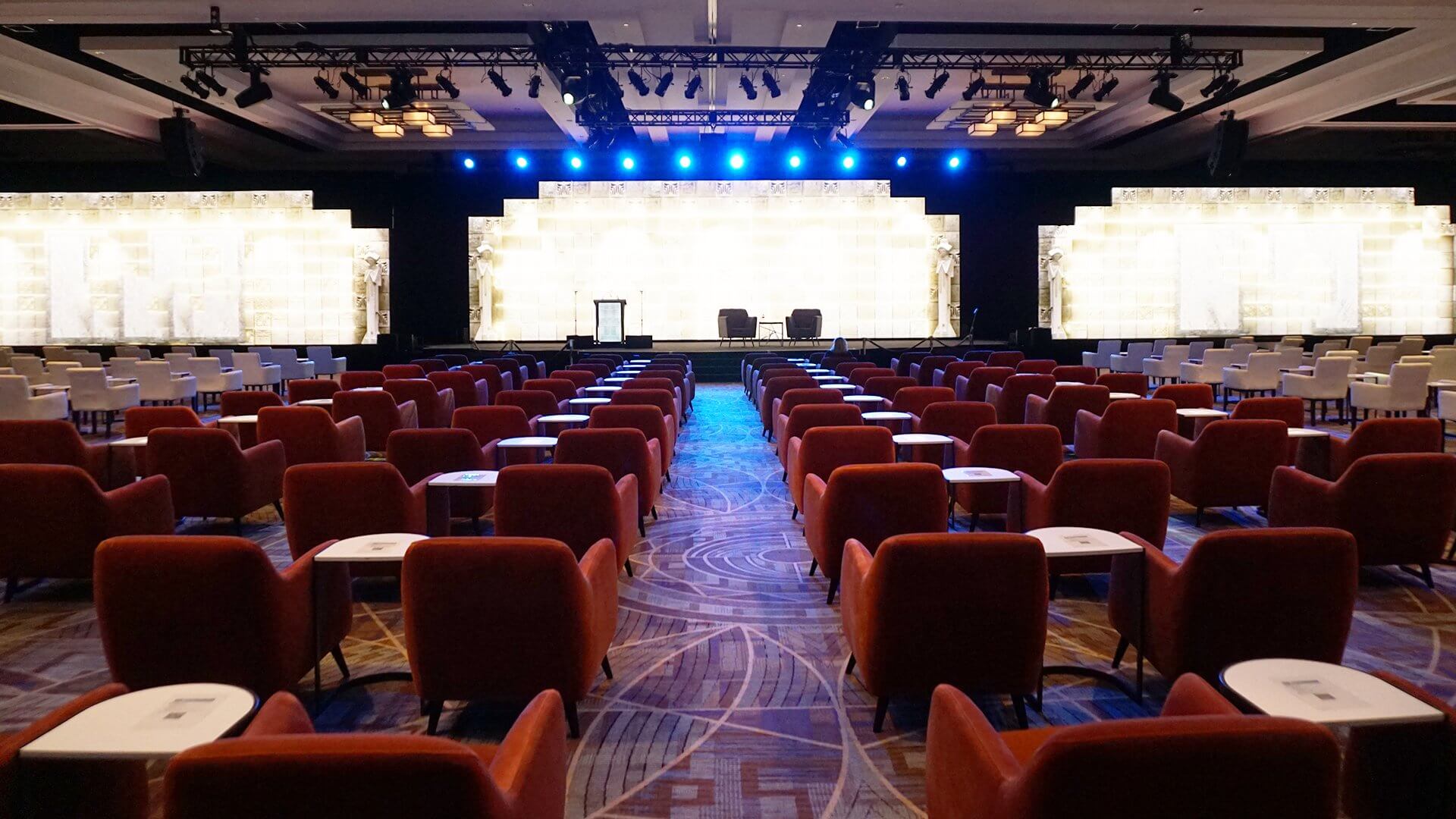 A large conference room is set up with rows of red chairs facing a brightly lit stage. The stage is adorned with decorative white panels and blue overhead lights. The space is ready for an event or presentation.