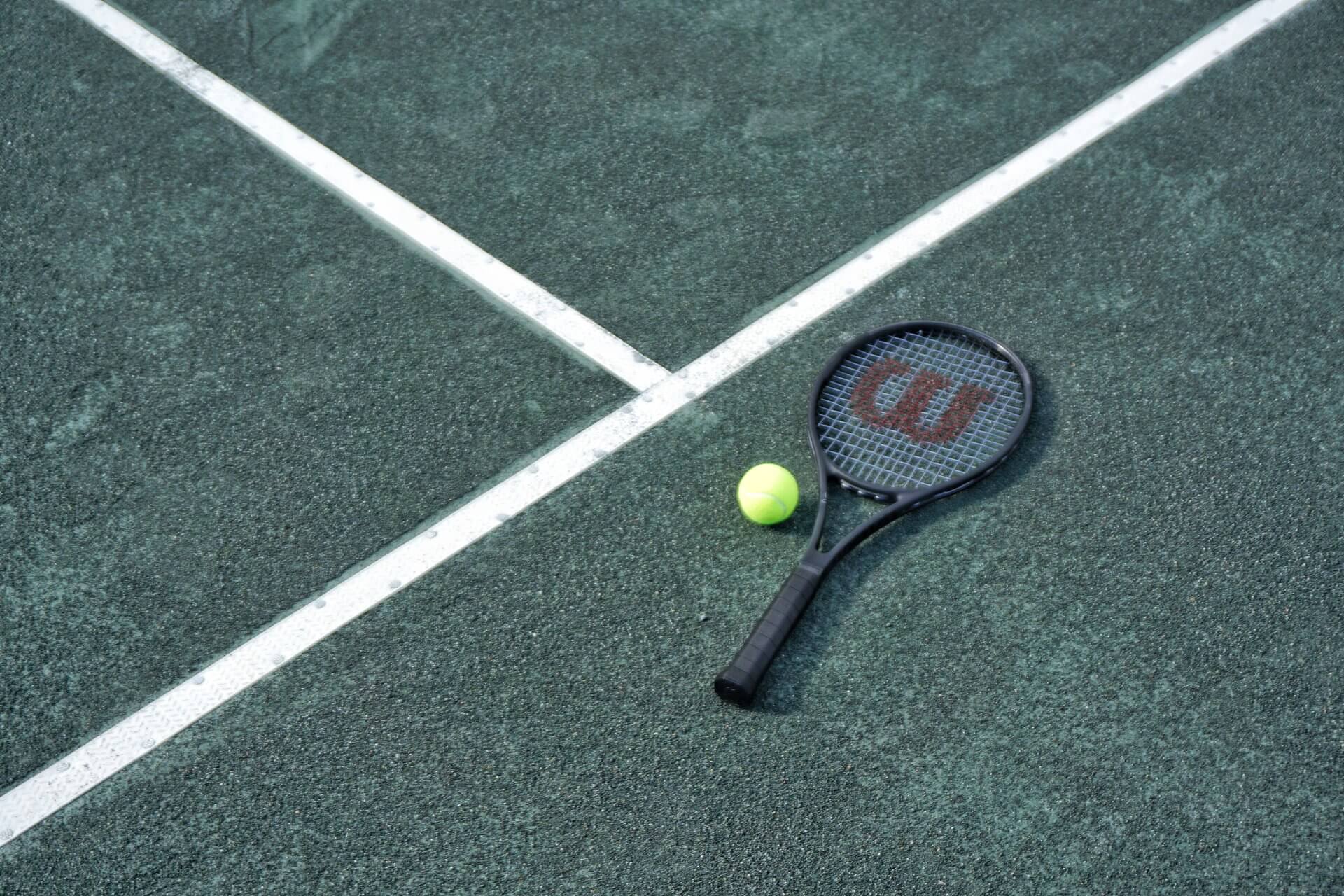 A tennis racket and a yellow tennis ball are positioned together on a green tennis court. The court's white boundary lines intersect near the racket and ball, creating a prominent cross shape in the background.