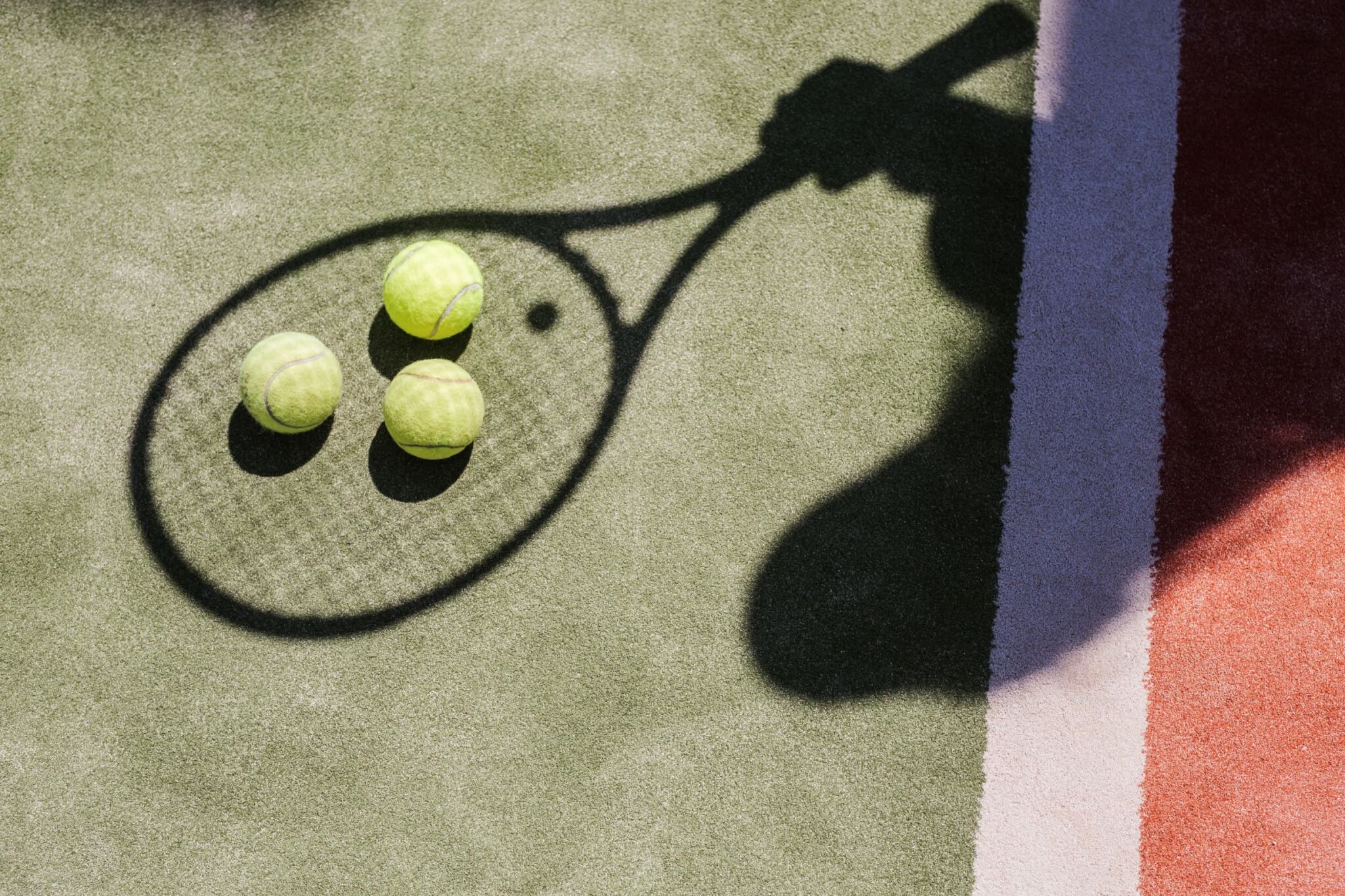 A tennis racket casts a shadow on the green textured surface of a tennis court, with three yellow tennis balls resting inside the shadow. The court features white and red boundary lines in the background.