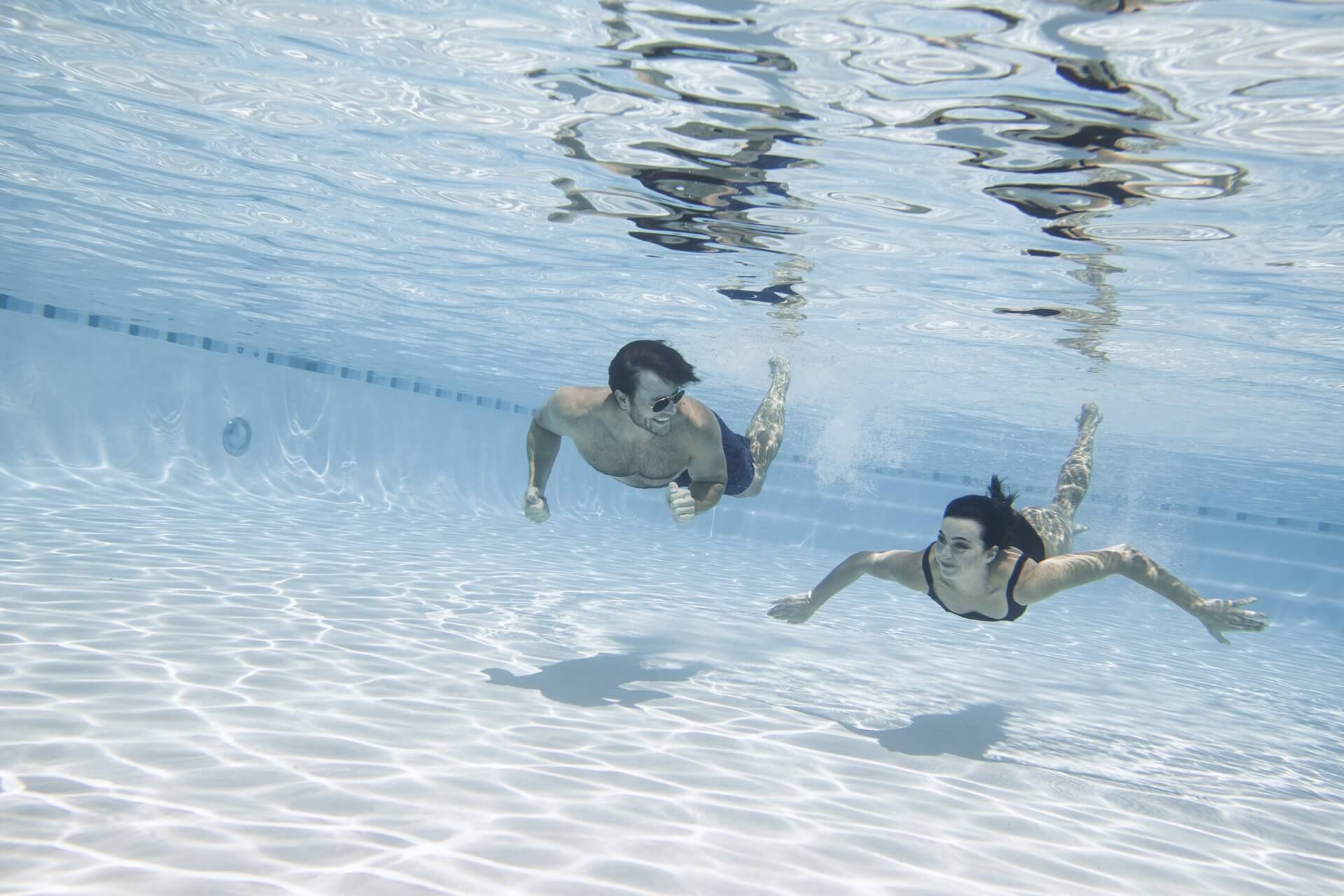 Two people, a man and a woman, are swimming underwater in a clear and well-lit pool. Both are wearing swimwear, with the man in trunks and the woman in a one-piece suit. The pool's tiled floor and clean water create a sparkling effect in the light, amplifying their enjoyment like it's 25% off happiness.