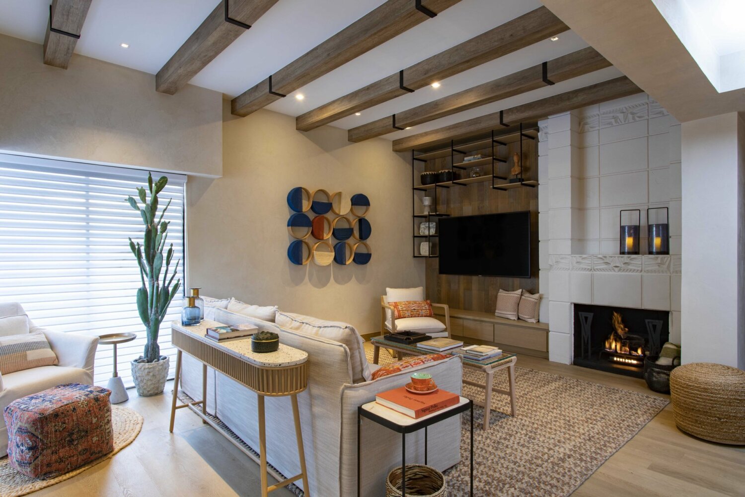A modern living room with wooden ceiling beams, neutral-toned furniture, and large windows with blinds invites you to stay. The space features a fireplace, a wall-mounted TV, a tall potted plant, and colorful wall art. Shelving units display decorative items, and there is a cozy area rug.