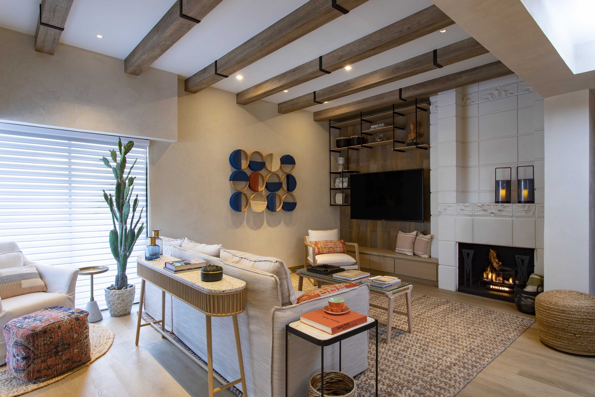 A modern living room with wooden ceiling beams, neutral-toned furniture, and large windows with blinds invites you to stay. The space features a fireplace, a wall-mounted TV, a tall potted plant, and colorful wall art. Shelving units display decorative items, and there is a cozy area rug.