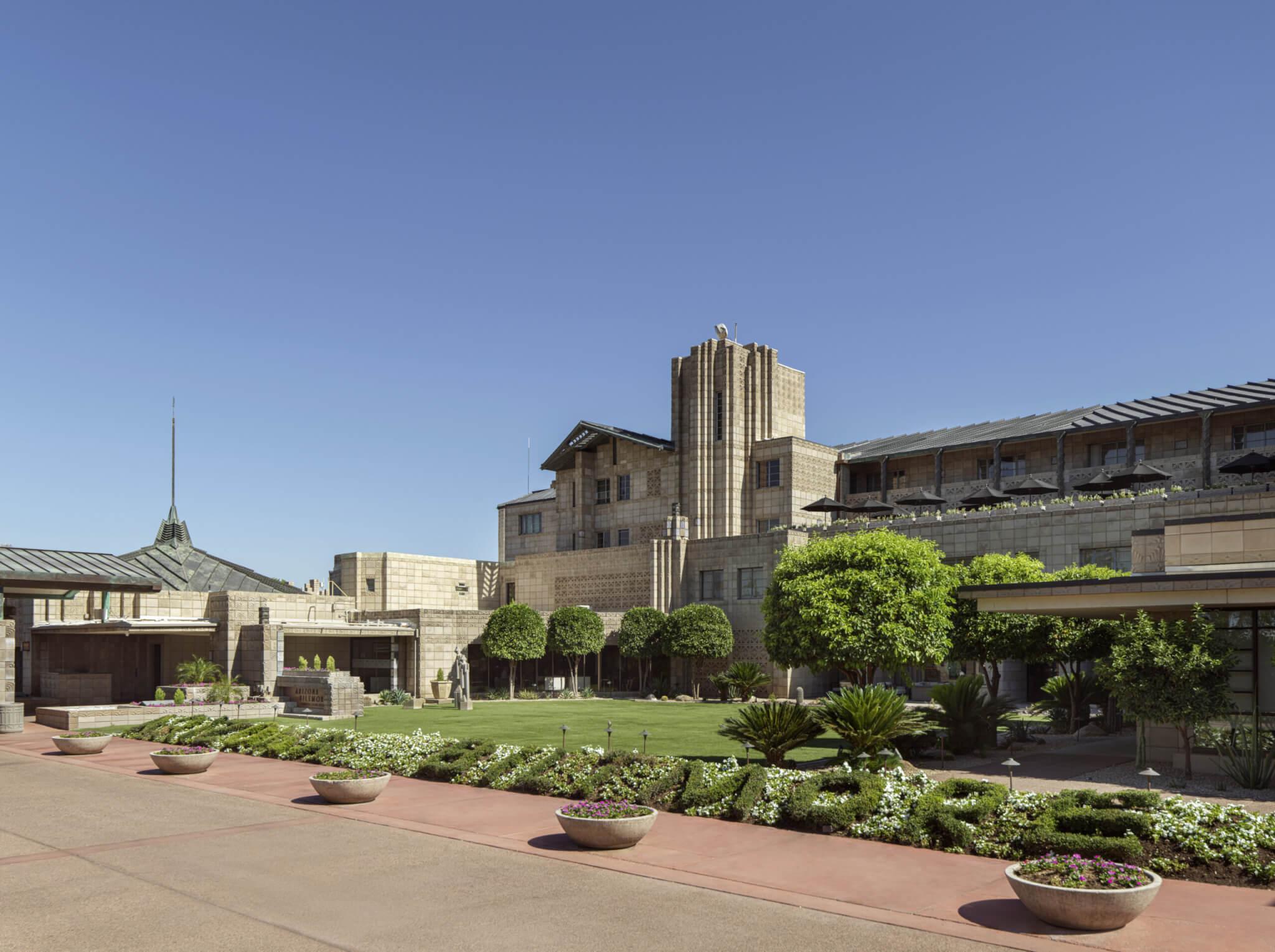 Exterior view of the historic Arizona Biltmore Hotel, a gem of our resort, designed by Frank Lloyd Wright. It showcases distinctive geometric patterns, lush landscaped gardens, and potted plants, all set against a clear blue sky.