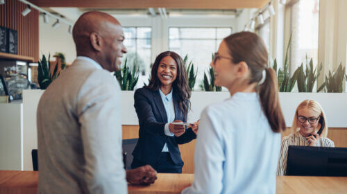 A smiling businesswoman standing behind a reception desk hands a card to a client, discussing potential careers. Another woman with glasses and a ponytail talks to the client, while a receptionist in the background works on a computer. The setting appears to be an office.