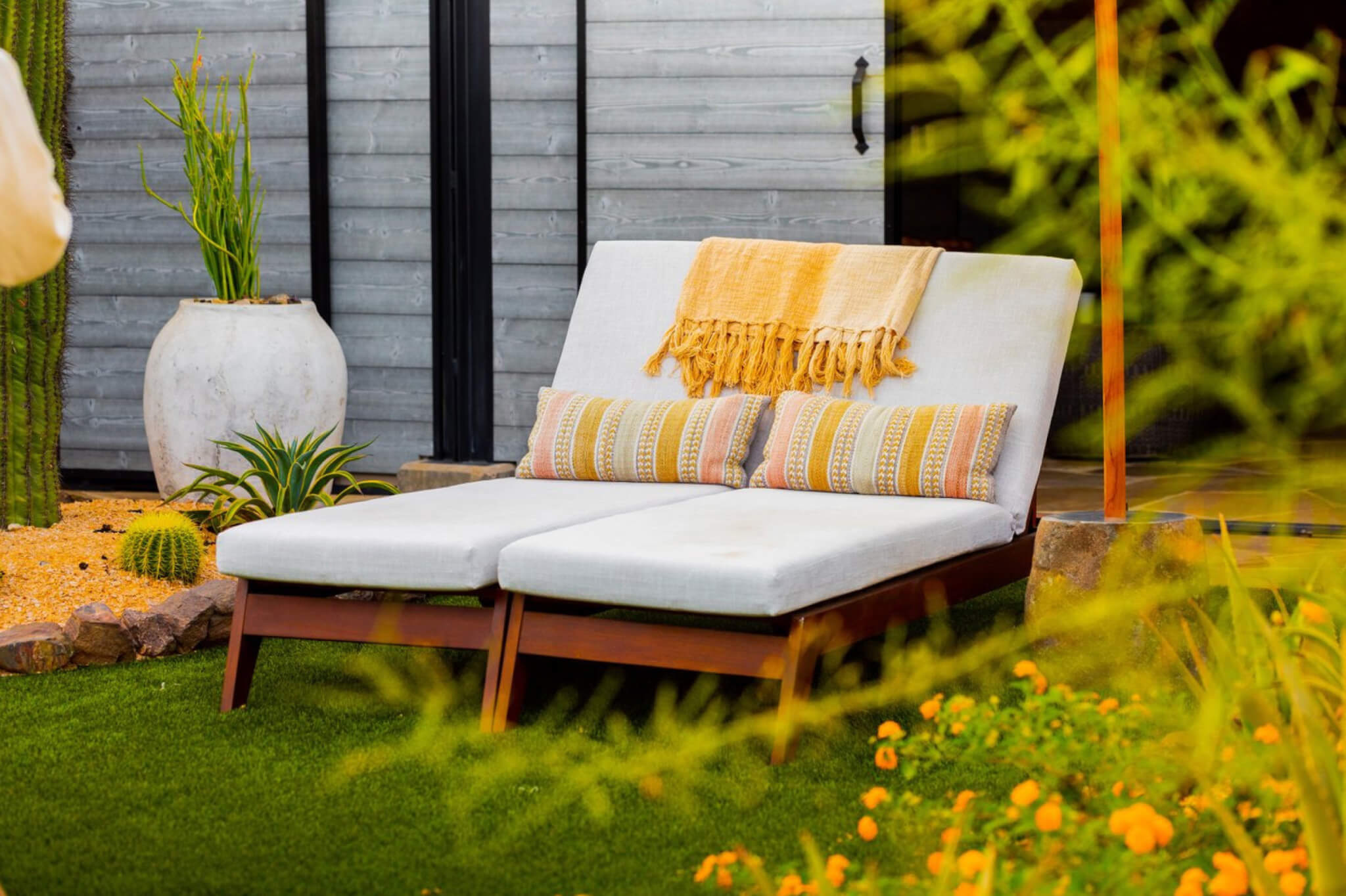 A serene outdoor setting reminiscent of Tierra Luna Spa featuring a cushioned double lounge chair with two patterned pillows and a yellow blanket, situated on a manicured lawn. The scene is enhanced by surrounding plants, a large ceramic planter, and a grey wooden wall in the background.