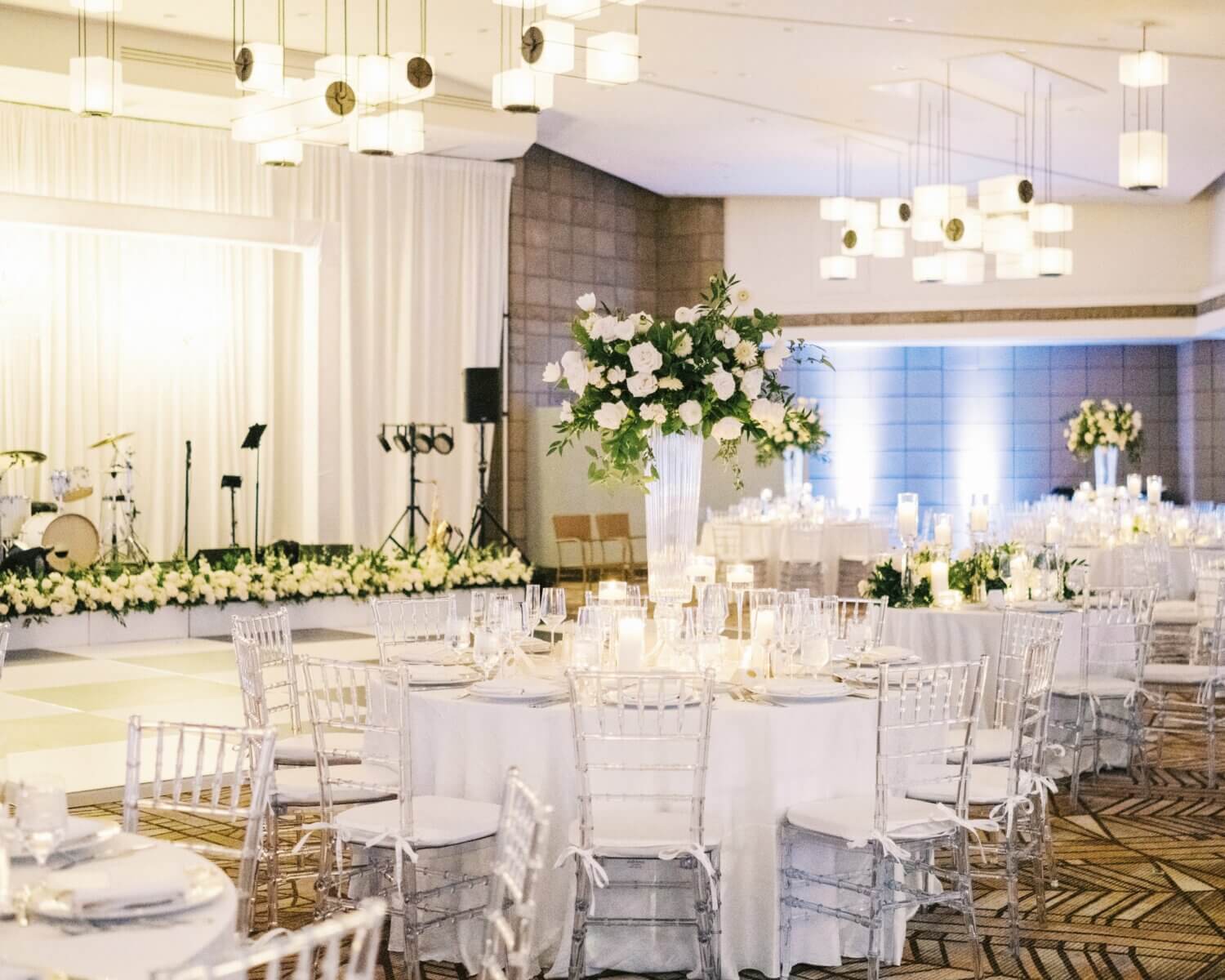 A beautifully decorated gold room reception hall with round tables covered in white linens and clear chairs. Each table is adorned with elegant centerpieces of white flowers. A stage in the background is lined with floral arrangements, with musical instruments set up nearby.