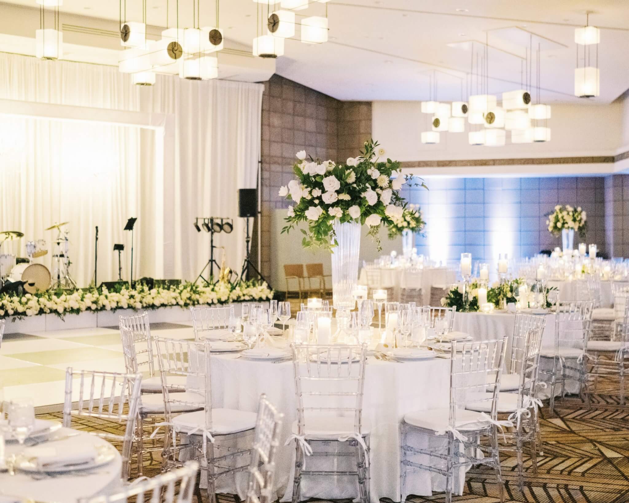 A beautifully decorated gold room reception hall with round tables covered in white linens and clear chairs. Each table is adorned with elegant centerpieces of white flowers. A stage in the background is lined with floral arrangements, with musical instruments set up nearby.