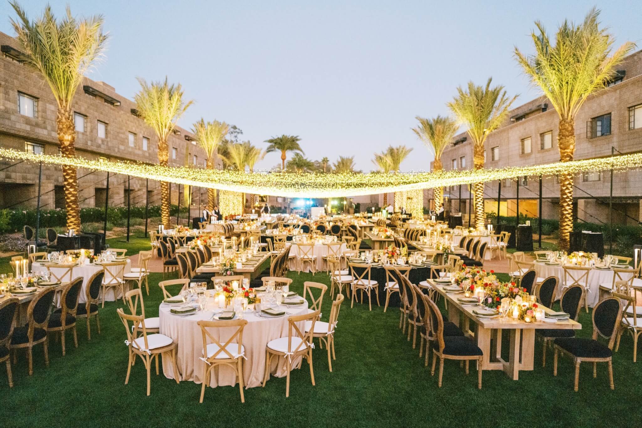 A beautifully decorated outdoor event setup during twilight with round and rectangular tables adorned with floral centerpieces, surrounded by chairs. Strings of fairy lights hung above create a cozy ambiance, transforming the setting into a paradise garden. Palm trees and buildings are visible in the background.