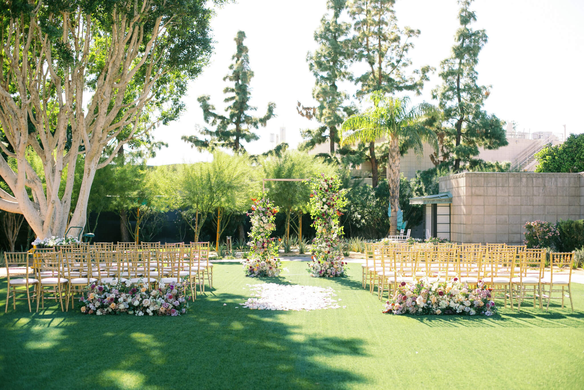 An outdoor wedding setup with rows of golden chairs arranged on a lush cottage lawn. The aisle, adorned with scattered white rose petals, leads to a floral arch surrounded by vibrant flower arrangements. Tall trees and bushes frame the serene backdrop.