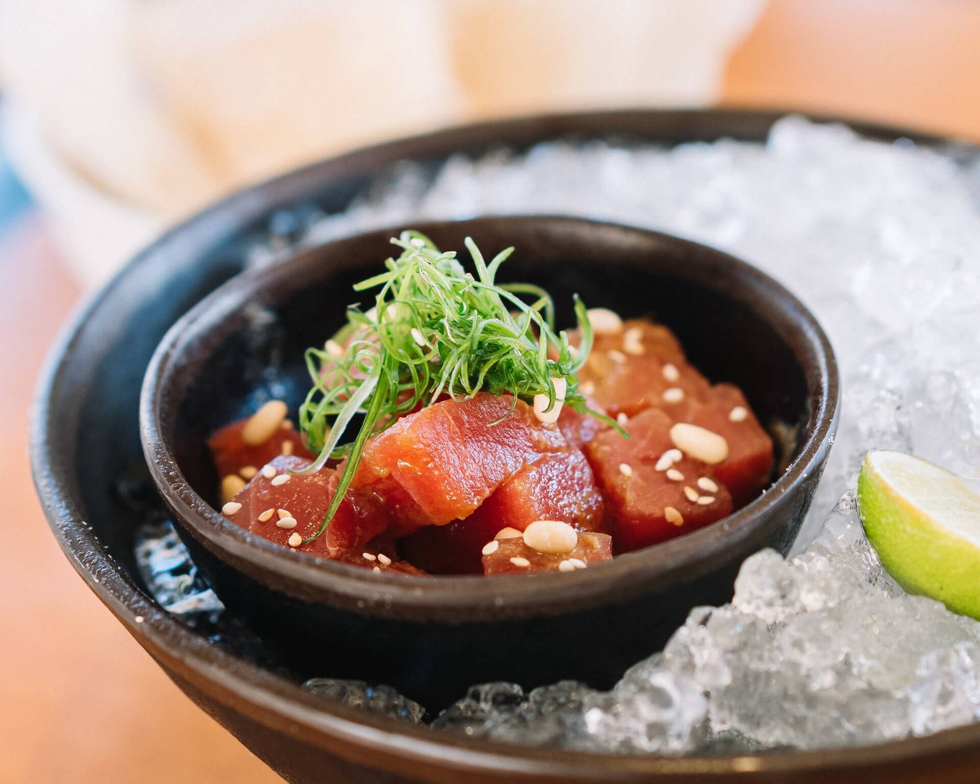 A close-up of a small black bowl filled with cubes of raw tuna garnished with chopped green onions and sesame seeds. The bowl is placed on a bed of ice in a larger black dish, reminiscent of a paradise garden, with a wedge of lime on the side.