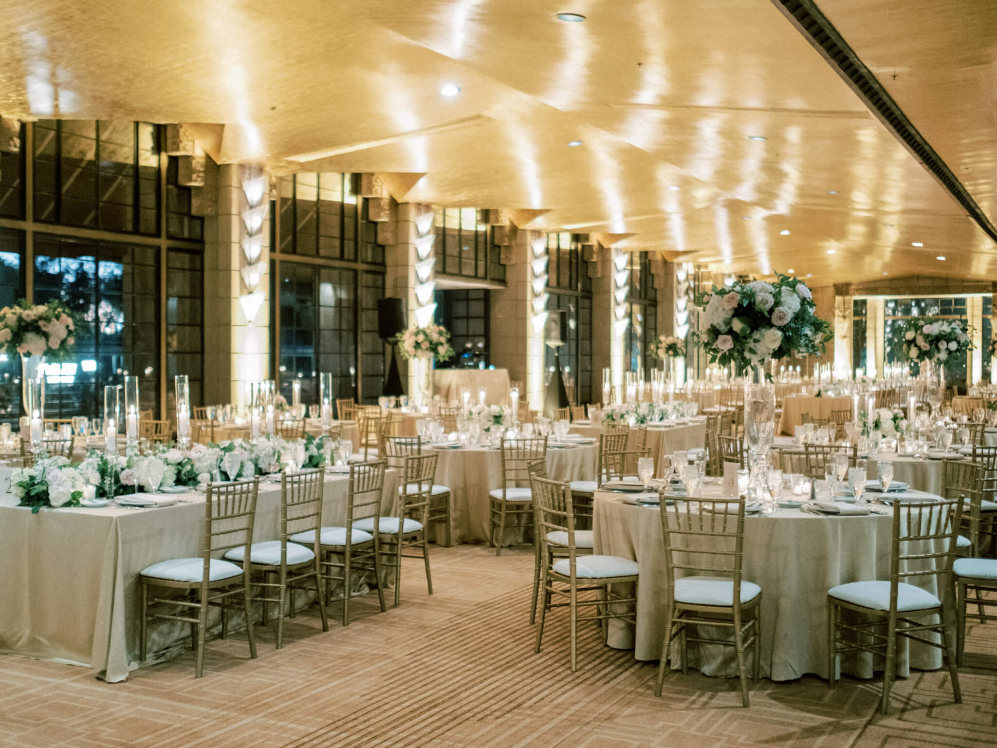 A large banquet hall, beautifully decorated for an event, features round tables covered in white linens, gold chiavari chairs, and elegant floral centerpieces. The gold room is softly lit with chandeliers and candles, creating a warm and inviting atmosphere.