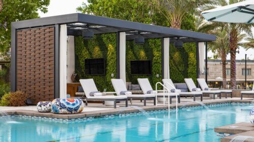 A modern outdoor lounge area beside the Saguaro pool features multiple white poolside chairs under a covered cabana. The cabana has lush green walls, two wall-mounted TVs, and decorative round cushions. The pool includes several water features and palm trees in the background.