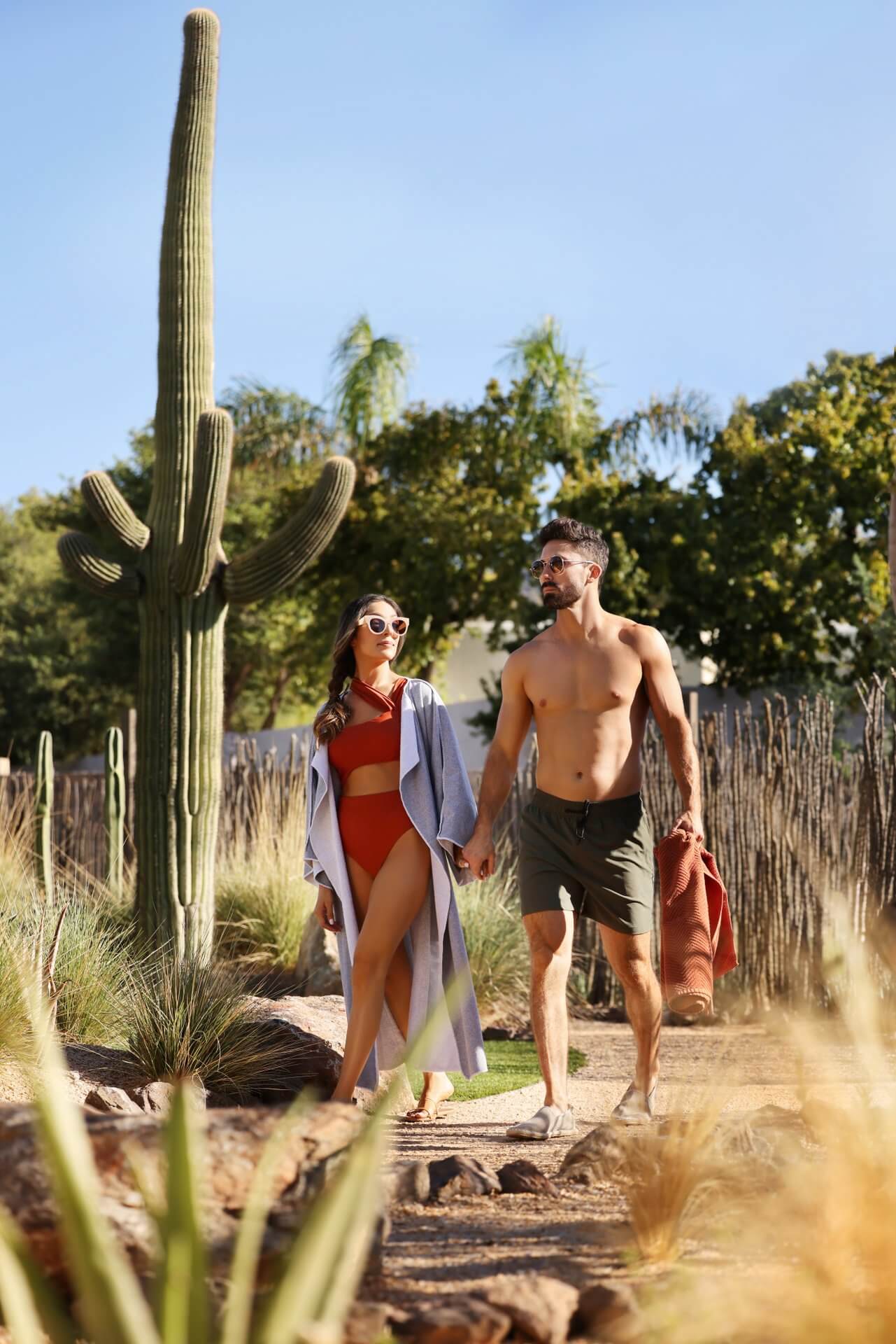 A couple walks hand in hand through a desert garden. The woman wears a red bikini and a light cover-up, while the man sports green swim trunks and holds a towel. Surrounded by various desert plants, including a tall cactus, they feel like they've found their own private Citrus Club under the bright, sunny sky.