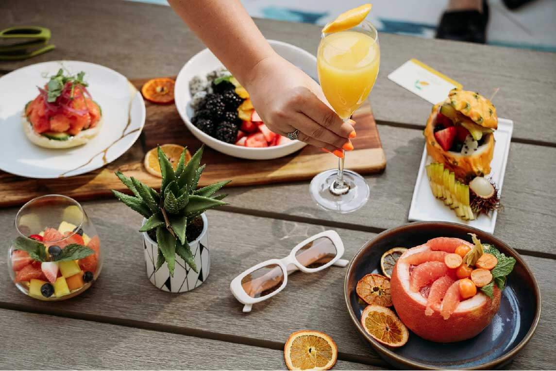 A brunch spread on a table features a colorful array of fresh fruits, a glass of citrus club orange juice, avocado toast with tomatoes, a bowl of mixed berries, and a pineapple filled with fruit salad. A potted plant and a pair of sunglasses complete the inviting scene.