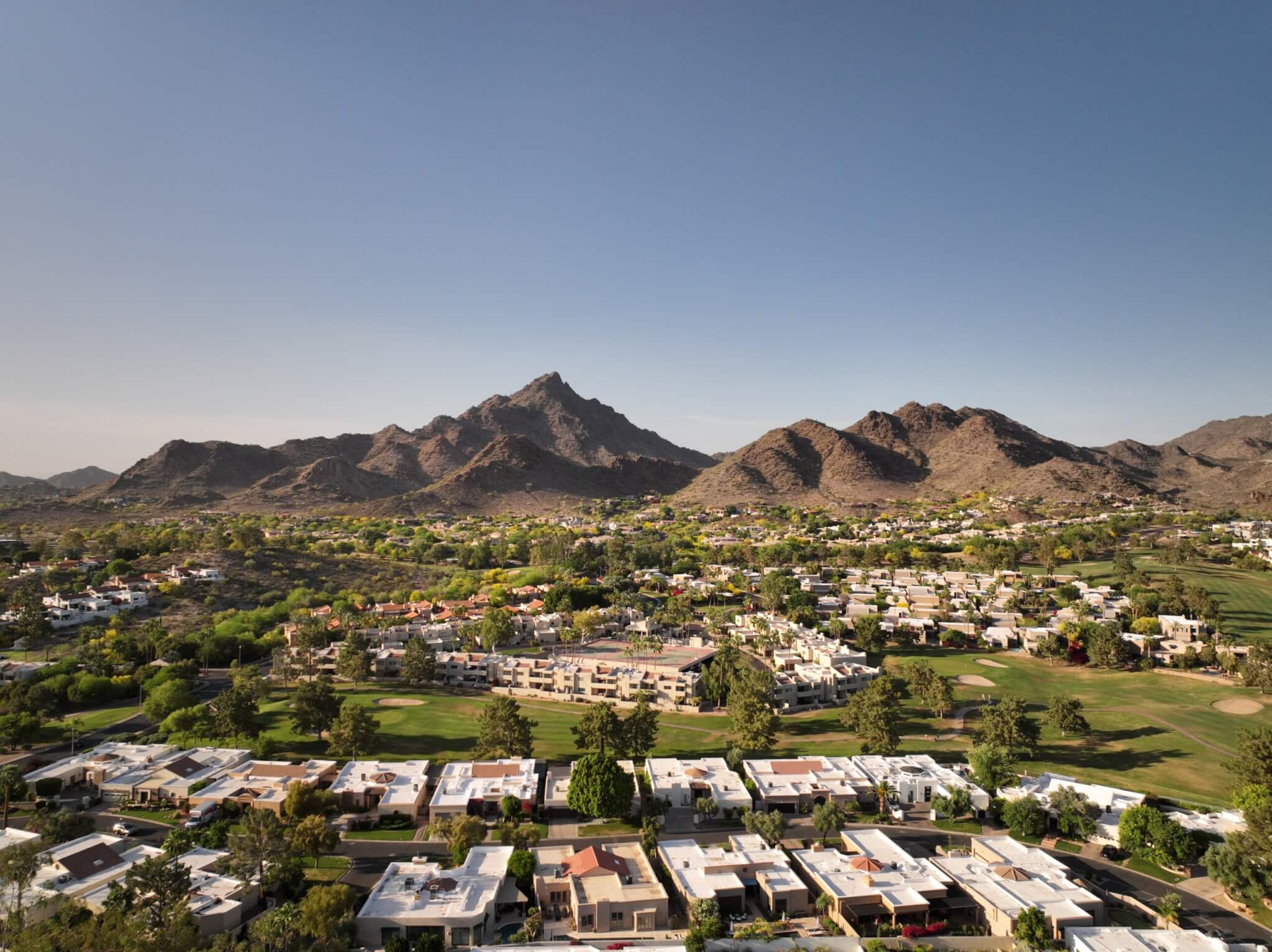 Aerial view of a suburban neighborhood with rows of houses, green lawns, and trees basking in the warm weather. In the background, rugged mountains under a clear blue sky dominate the landscape. The overall scenery is tranquil and picturesque.