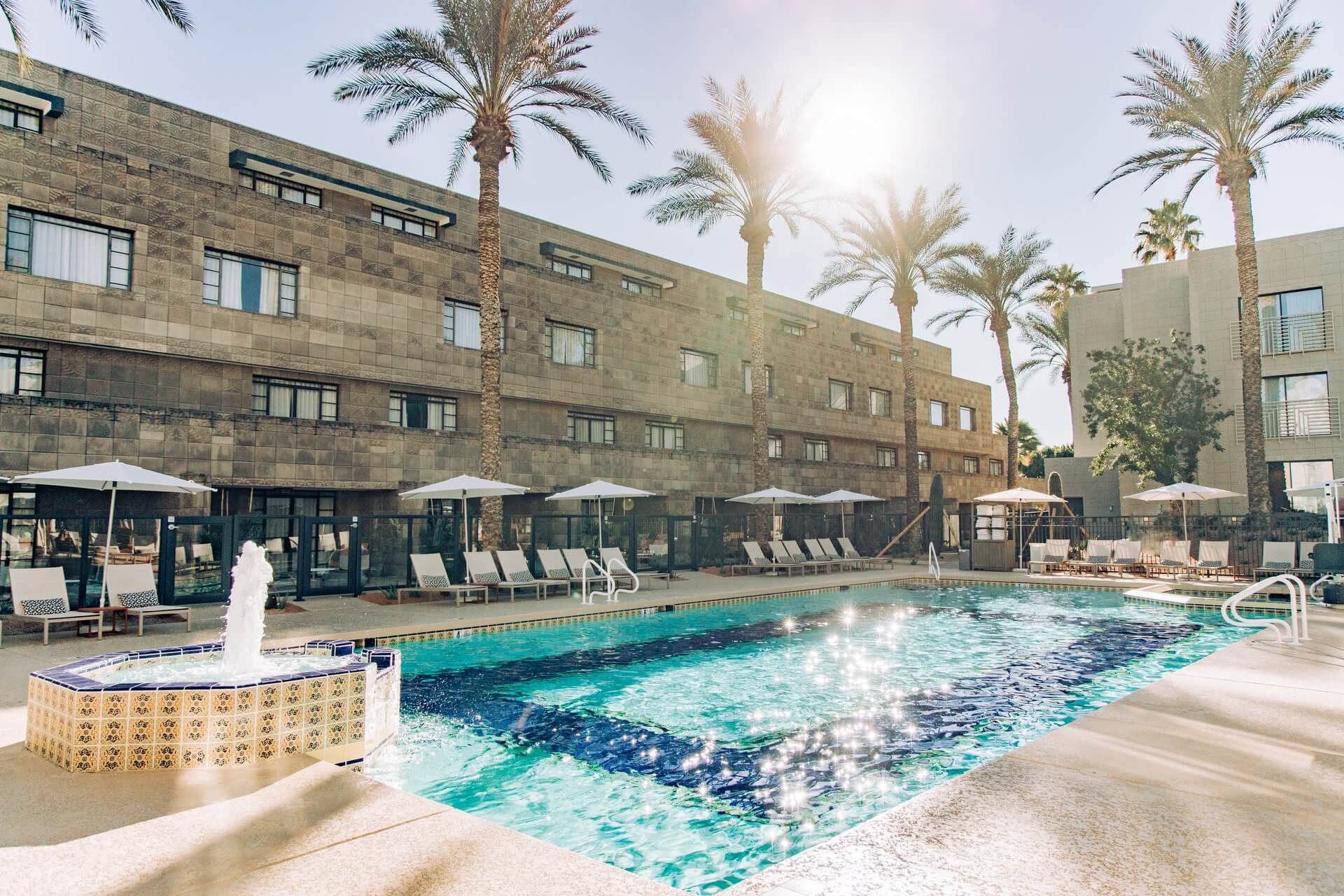A serene outdoor swimming pool is surrounded by lounge chairs and umbrellas. Palm trees stand tall around the area, and sun rays illuminate the scene. In the background, a modern brick building adds to the charm of one of those luxurious resort pools. A small fountain is located at one end of the pool.