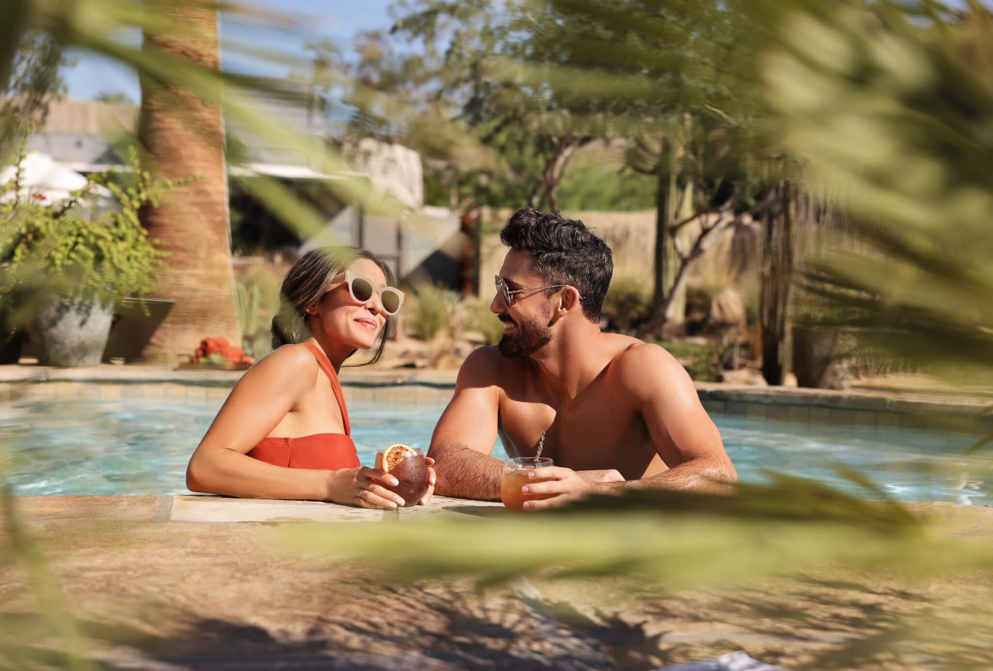 A woman and a man are smiling at each other while relaxing in a pool under the summer sunlight. The woman in a red swimsuit holds a drink and an orange slice. Both are wearing sunglasses. Palm fronds frame the foreground, giving a tropical ambiance.