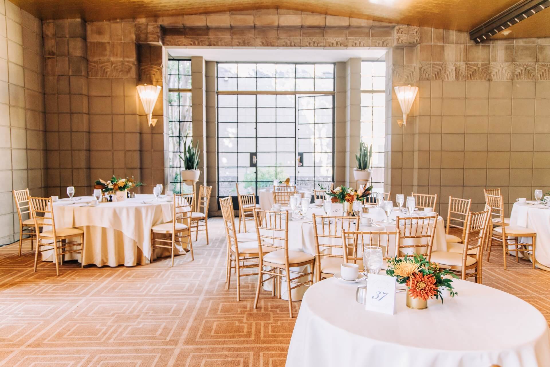 A well-lit banquet hall with round tables covered in white tablecloths. Each table is adorned with flower centerpieces and surrounded by wooden chairs. The room, perfect for an Easter celebration, features large windows, soft lighting from wall sconces, and potted plants by the windows.