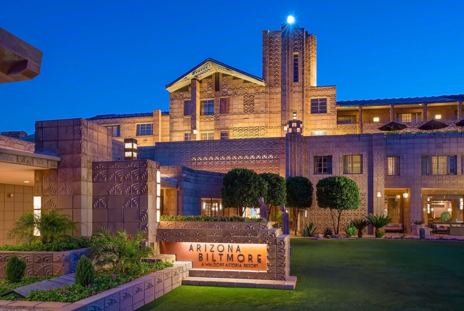 A view of the Arizona Biltmore resort at dusk, showcasing its grand architecture illuminated with lights. The main entrance features a sign reading "Arizona Biltmore," surrounded by manicured lawns, trees, and ornamental designs on the building facade—perfect for a summer evening stroll.