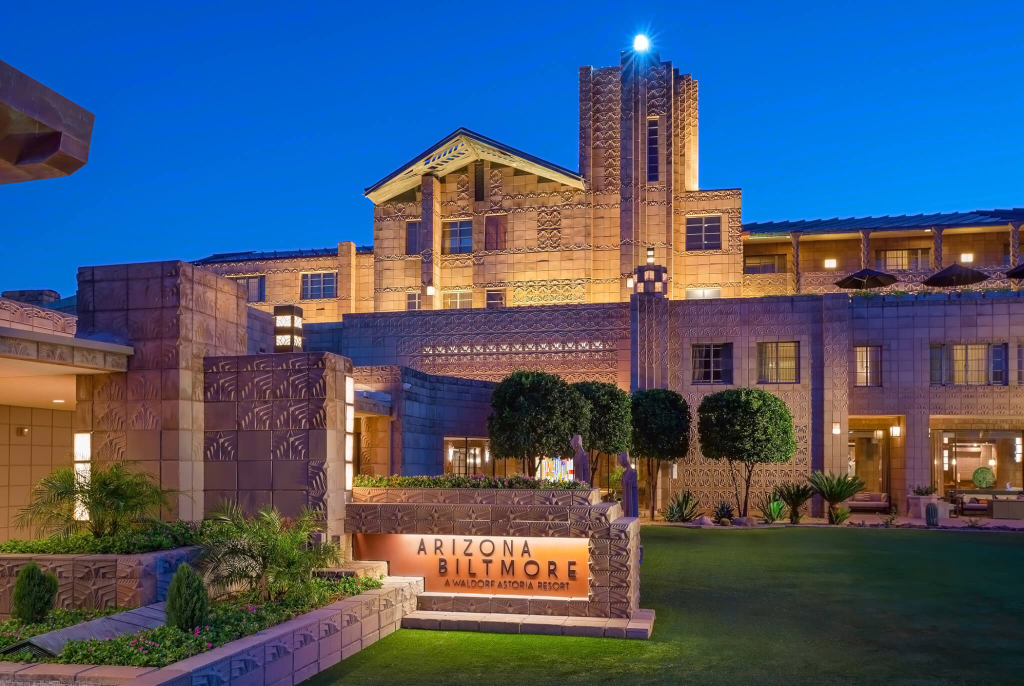 A view of the Arizona Biltmore resort at dusk, showcasing its grand architecture illuminated with lights. The main entrance features a sign reading "Arizona Biltmore," surrounded by manicured lawns, trees, and ornamental designs on the building facade—perfect for a summer evening stroll.