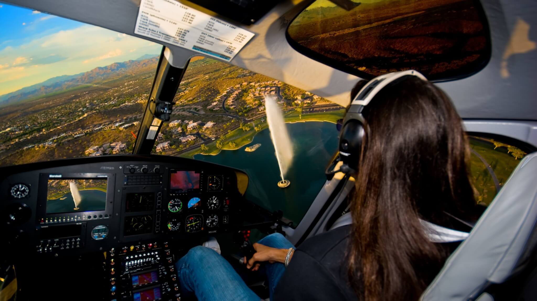 A person with long hair wearing headphones pilots a helicopter over a city, flying above a large lake with a tall fountain. The Amethyst Expedition unfolds as the helicopter’s dashboard and scenic landscape are visible through the windows.